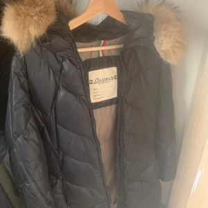 Svart dunjacka med päls  från Dolomite - Svart dunjacka från Dolomite med stor fluffig fuskpäls på huvan och snygga quiltade detaljer. Jackan har dragkedja framtill och en badge på ärmen. Perfekt för kalla dagar när du vill hålla stilen och värmen.