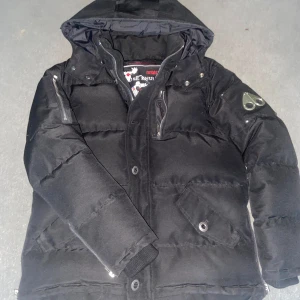 Svart Moose Knuckles jacka - Svart dunjacka från Moose Knuckles med huva, dragkedjor på ärmarna och snygga detaljer som logopatch på axeln. Jackan har broderad insida med hockeymotiv och flera fickor framtill. Perfekt för kalla dagar när du vill hålla stilen. Zippern är tyvärr sönder men inget märk med knapparna på