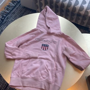 Rosa hoodie från GANT med logga - Snygg ljusrosa hoodie från GANT med broderad logga och emblem på bröstet. Tröjan har huva, magficka och ribbade muddar. Perfekt för chill dagar och har en klassisk, sportig vibe. Materialet är mjuk bomull som känns skönt mot huden.