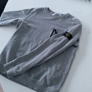 Grå sweatshirt från Stone Island Junior - Grå sweatshirt från Stone Island Junior med klassisk rund hals och lång ärm. Tröjan har en svart dragkedja på bröstfickan och den ikoniska Stone Island-patchen på vänster ärm. Perfekt för dig som gillar stilrena och sportiga plagg.