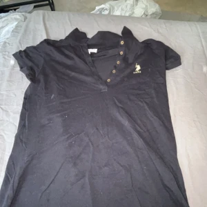 Svart polotopp från US Polo Assn M - Svart polotopp från US Polo Assn med klassisk krage och knappslå med fem metallknappar. Toppens material är mjuk bomull och har en broderad logga i guld på bröstet. Perfekt för dig som gillar sportig och stilren vibe.