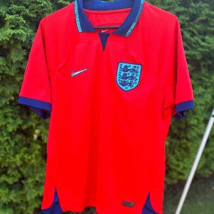 England röd fotbollströja Nike - Röd England fotbollströja från Nike med blå detaljer och broderat landslagsmärke på bröstet. Tröjan har korta ärmar, blå krage med ljusblå ränder och är tillverkad i ett lätt, ventilerande material perfekt för match eller träning. Har ni fler frågor om lappar eller annat är det bara att höra av sig! Priset är ej hugget i sten👍