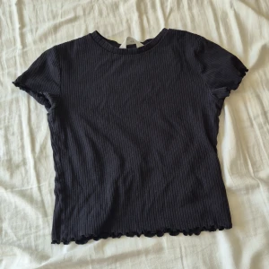 Svart ribbad topp från H&M - En svart ribbad topp från H&M med korta ärmar och volangkant nertill. Toppen har en rund halsringning och är i stretchigt material som sitter snyggt på kroppen. Perfekt för dig som gillar enkel men trendig stil.