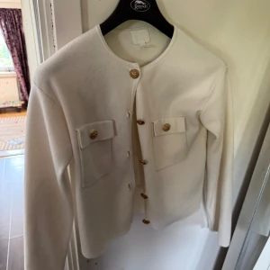 Vit kofta med guldknappar från H&M - Supersnygg vit kofta från H&M med rund hals och dekorativa guldknappar framtill. Två stora fickor med lock och knappar ger en cool touch. Mjuk och skön, perfekt för lager-på-lager. Långärmad och i ett stilrent snitt som funkar till allt.