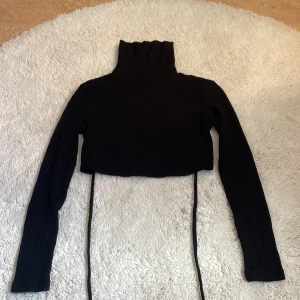 Svart croppad Bershka med snörning - Säljer en svart croppad Bershka tröja med långa ärmar och ribbad struktur. Tröjan har snygg snörning som knyts runt midjan för en trendig look. Perfekt att styla med höga jeans eller mjukisbyxor. Materialet är mjukt och stretchigt, vilket gör den bekväm att bära.