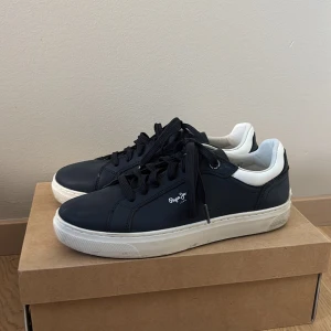 Mörkblå sneakers från Pepe Jeans - Snygga mörkblå sneakers från Pepe Jeans med vita detaljer och bruna gummisulor. Skorna har snörning, rund tå och texten '1973' i guld på hälen. Pepe Jeans-logga på sidan och undersidan. Perfekt för dig som gillar stilrena och klassiska sneakers.