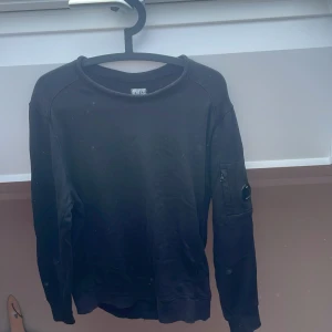 Svart sweatshirt från C.P. Company - Svart långärmad sweatshirt från C.P. Company med rund hals och ikonisk linsdetalj på vänster ärm. Tröjan har en enkel, clean look och är tillverkad i mjuk bomull. Perfekt för dig som gillar streetwear och vill ha något stilrent med cool detalj.