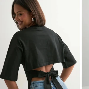 Svart croppad topp med knyt i ryggen - Svart croppad topp med halvlånga ärmar och öppen rygg som knyts med en stor rosett baktill. Snygg och trendig modell som passar perfekt till jeans eller kjol. Toppens material ser ut att vara bomull eller liknande för en skön känsla.