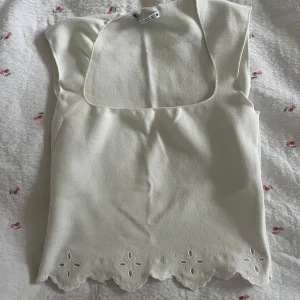 Vit linnetopp med broderad spetskant - Supersöt vit linnetopp från Zara med fyrkantig ringning och broderad spetskant nertill. Ärmlös modell i mjukt tyg, perfekt för varma dagar. Blommiga broderidetaljer längs nederkanten ger en romantisk vibe. Storlek M men är liten i storlek!!
