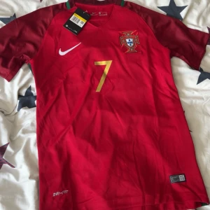 Portugal Ronaldo 7 Nike fotbollströja  - Portugal landslagströja från Nike med Ronaldo 7-tryck i guld på ryggen. Röd bas med mörkröda ärmar, vita och guldiga detaljer samt broderat landslagsmärke. Tillverkad i Dri-FIT-material som andas, perfekt för fotboll eller träning.