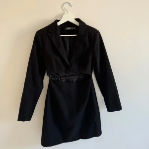 Svart cut-out blazerklänning  - Svart kort kostymklänning från Missguided Petite med snygg cut-out detalj i midjan. Klänningen har lång ärm, klassisk krage och knäppning framtill. Perfekt för dig som gillar trendiga och edgy looks. Knapp fram. Dragkedja i sidan