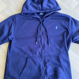 Marinblå hoodie från Polo Ralph Lauren - Säljer nu min Polo Ralph lauren Zip hoodie som ej kommer till användning, köptes i våras men har ej används något bara testad, tröjan har inga som helst tecken på användning och är riktigt stilren och snygg. Perfekt för dig som gillar det lilla extra, vid frågor angående plagget tveka inte kontakta mig vänliga hälsningar!