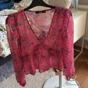 Blommig volangblus från BikBok XS - Superfin blommig blus från BikBok i transparent rosa och lila med volangdetaljer och v-ringning. Långärmad och luftig modell som ger en romantisk vibe. Perfekt för dig som gillar färg och mönster!