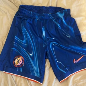 Chelsea x Nike blå fotbollsshorts Storlek M - Snygga blå Chelsea fotbollsshorts från Nike med coolt mönster i olika blå toner och orange detaljer. Shortsen har elastisk midja, Chelsea FC-logga och orange Nike Swoosh. Tillverkade i Dri-FIT polyester och har slim fit passform. Perfekta för fotbollsträning eller match.