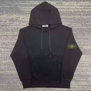 Svart hoodie från Stone Island - Svart hoodie från Stone Island med klassisk logopatch på vänster ärm. Tröjan har huva med snörning och en stor magficka framtill. Perfekt för dig som gillar streetwear och vill ha en clean look med snygga detaljer.