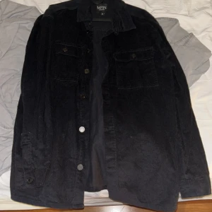 Svart manchester overshirt BoohooMAN - Säljer en svart overshirt från BoohooMAN i mjuk manchester. Skjortan har klassisk krage, knäppning framtill och två bröstfickor med lock. Perfekt lager-på-lager-plagg med avslappnad passform och snygg struktur.