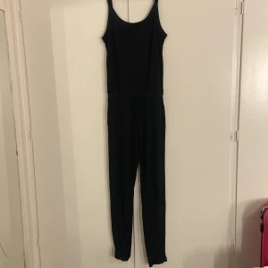 Svart jumpsuit från Hunkemöller - Svart ribbad jumpsuit från Hunkemöller i storlek S. Modellen har smala axelband, rundad halsringning och snörning i midjan för en snygg passform. Materialet är mjukt och stretchigt, perfekt för chill dagar eller när du vill vara bekväm.