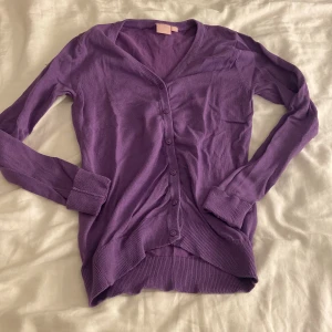 Lila kofta med v-ringning - Säljer en lila kofta med v-ringad hals och långa ärmar. Koftan har knappar hela vägen framtill och ribbade muddar vid ärmslut och nederkant. Perfekt att slänga över en t-shirt för en chill look.