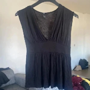 Svart topp från H&M - Snygg svart topp från H&M med djup v-ringning och bred midjedetalj. Toppen är ärmlös och har ett lätt, mjukt tyg som faller fint. Perfekt för dig som gillar enkel men stilren design.