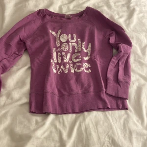 Lila sweatshirt med tryck - Säljer en lila sweatshirt med vitt tryck på framsidan där det står 'You only live twice'. Tröjan har långa ärmar, rund hals och breda muddar vid ärmslut och nederkant. Perfekt för chill dagar och enkel att matcha med jeans eller leggings.