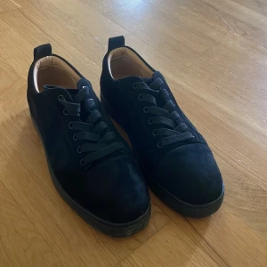 Svarta Louboutin  - Snygga svarta sneakers i mocka från Christian Louboutin med klassiska röda sulor och guldfärgad logga på innersulan. Skorna har rund tå, svarta skosnören och diskreta detaljer. Perfekta för dig som vill ha en lyxig och stilren look.
