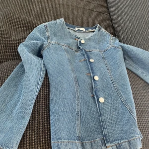 Ljusblå jeansjacka med knappar - Säljer en stilren ljusblå jeansjacka med rund halsringning och silverfärgade knappar framtill. Jackan har långa ärmar och klassiska sömmar som ger en snygg passform. Perfekt för dig som gillar en clean och tidlös look.