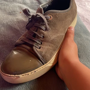 Lanvin sneakers i mocka och läder - Säljer ett par snygga Lanvin sneakers i mörkbrun mocka med läderdetaljer och blank tå. Skorna har rund tå, snörning och beige platt sula. Perfekt för dig som gillar stilrena och lyxiga sneakers med unik design.