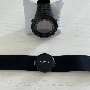 Svart sportklocka Suunto med pulsbälte - Sportig svart Suunto-klocka med digital display och robust silikonarmband. Medföljer matchande svart pulsbälte för träning och pulsmätning. Perfekt för dig som gillar att hålla koll på din aktivitet och vill ha en stilren och funktionell accessoar.