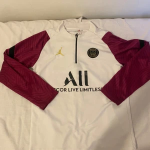 PSG x Jordan långärmad fotbollströja M - Snygg PSG fotbollströja i samarbete med Jordan. Vit bas med vinröda, mönstrade ärmar och svarta detaljer. Halvlång dragkedja framtill, guldigt Jordan-logga och PSG-märke på bröstet. Tillverkad i lätt och ventilerande material, perfekt för träning eller match.