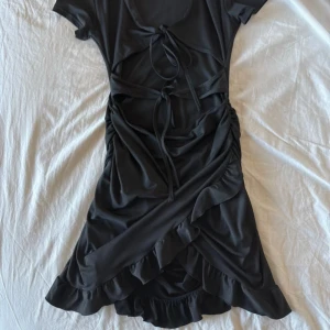 Svart omlottklänning från SHEIN - Supersnygg svart kortklänning från SHEIN med omlott-design, volangkant och coola snörningsdetaljer framtill. Klänningen har korta ärmar och är gjord i mjukt polyester-material som sitter skönt mot kroppen. Perfekt för dig som gillar trendiga och unika plagg!