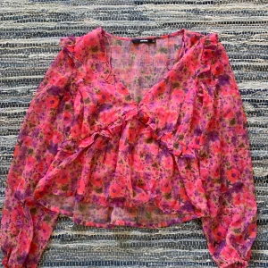 Blommig volangblus från BikBok XS - Supersöt blommig blus från BikBok i tunnt, transparent material. Färgerna går i rosa, lila och grönt med volangdetaljer och v-ringning. Långärmad modell med lätt puff vid axlarna och resår vid ärmslut. Perfekt för dig som gillar färg och romantiska vibes!