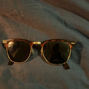 Ray-Ban Clubmaster solglasögon brun/guld - Snygga Ray-Ban Clubmaster solglasögon med klassisk brun sköldpaddsmönstrad ovandel och guldfärgade detaljer. Glasen är mörka och bågarna har den ikoniska retrolooken. Perfekta för dig som vill ha en tidlös och cool stil.