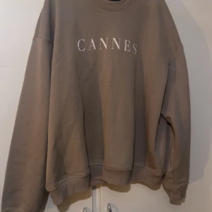 Beige sweatshirt med CANNES-tryck - Säljer en oversized beige sweatshirt med trycket 'CANNES' i vitt på bröstet. Tröjan har rund hals, långa ärmar och ribbade muddar vid ärmslut och nederkant. Perfekt för chill dagar och enkel att matcha med jeans eller leggings.