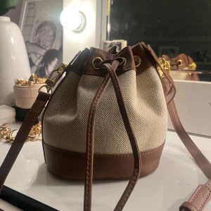 Beige och brun bucket bag med snörning - Säljer en snygg bucket bag i beige canvas med bruna skinn-detaljer och guldfärgade metalldelar. Väskan har justerbar axelrem och stängs med snörning upptill. Perfekt storlek för det viktigaste och ger en chill men ändå stilren vibe.