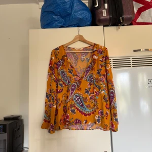 Gul paisleymönstrad blus med v-ringning - Färgstark blus från Odd Molly i en mörkgult/ orange färg med paisleymönster i blått, vinrött och rosa. Blusen har v-ringning, långa ärmar och en lös passform. Perfekt för dig som gillar boho-vibes och vill sticka ut med coola färger och mönster. Storlek 3 som enligt deras storleksguide motsvarar en storlek L.  Mycket fint skick, jag hittar inga synliga anmärkningar eller defekter.