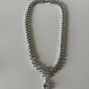 Glittrigt halsband med stora stenar - Halsband i silverfärg med massor av gnistrande stenar och en stor droppformad kristall som hänge. Kedjan är täckt av små, glittrande stenar i olika former.