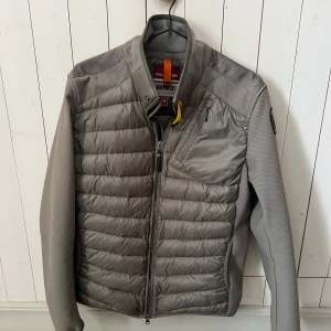 Grå pufferjacka från Parajumpers M - Grå pufferjacka från Parajumpers med ärmar i polyester. Jackan har dragkedja, bröstficka med dragkedja och den klassiska gula detaljen vid halsen. Har prislappen som hängde på den när den köptes och onlinekvitto från johnells. Kom gärna med bud!