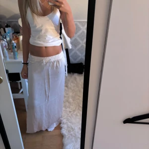 Vit långkjol med slits och snörning - Säljer en vit långkjol med elastisk midja och snörning. Kjolen har en snygg slits på sidan som ger en chill vibe och är gjord i ett tunt, flowy material som passar perfekt till varma dagar.