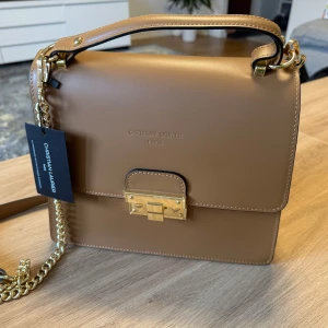 Helt ny väska – aldrig använd!Beige axelväska från Christian Laurier - Snygg beige axelväska från Christian Laurier med stilren design och guldfärgade metalldetaljer. Väskan har ett elegant lås framtill, kedjerem och ett praktiskt handtag upptill. Tillverkad i slätt skinn som ger en lyxig känsla. Perfekt för dig som gillar klassiska accessoarer.Väskan kom när jag var utomlands och jag hann inte returnera den i tid. Därför säljer jag den nu till ett mycket lägre pris. Perfekt för dig som vill ha en helt ny väska till ett riktigt bra pris!