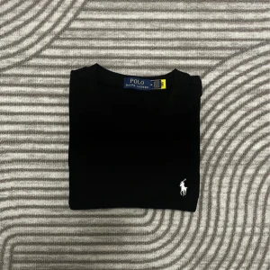 Svart t-shirt från Polo Ralph Lauren - Svart t-shirt från Polo Ralph Lauren med klassisk rund hals och korta ärmar. På bröstet finns den ikoniska vita Polo-loggan broderad. T-shirten är gjord i mjuk bomull och har en clean, stilren look som funkar till allt.