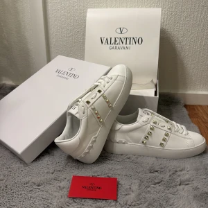 Valentino Rockstud vita sneakers strl 38 - Snygga vita Valentino Rockstud sneakers i skinn med ikoniska nitar i guld längs sidorna och bak på sulan. Klassisk rund tå, snörning och platt sula. Perfekta för dig som vill ha en lyxig och trendig look med edge.