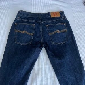 Mörkblå jeans från Nudie Jeans, 32/32 - Säljer ett par mörkblå jeans från Nudie Jeans i klassisk modell med snygga kontrastsömmar och ikonisk brodyr på bakfickorna. Jeansen har normal passform och är tillverkade i slitstarkt bomullsjeans. Perfekta för dig som gillar stilrena och tidlösa jeans.