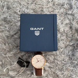 Guldbrun klocka från GANT med läderband - Stilren klocka från GANT med vit urtavla, guldfärgad boett och brunt läderband med krokodilmönster. Urtavlan har minimalistiska index och en extra liten visare för datum. Klockan levereras i originalask och har detaljer i rostfritt stål.