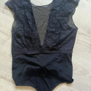 Svart spetsbody med djup v-ringning - Snygg svart body med djup v-ringning och spetsdetaljer upptill. Överdelen har transparent mesh och vackert blommönstrad spets, medan nedre delen är i slät, stretchig syntet. Från pieces, storlek xs