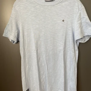 Ljusblå t-shirt från Morris XS - Snygg ljusblå t-shirt från Morris i storlek XS. T-shirten har rund hals, korta ärmar och en liten Morris-logga broderad på bröstet. Materialet är mjuk bomull och nederkanten pryds av en exklusiv Morris-patch. Perfekt för dig som gillar stilrena plagg med diskreta detaljer.