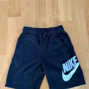 Svarta Nike shorts med logga 147-158cm - Nike shorts i svart med stor vit logga på ena benet. Modellen har elastisk midja med snörning och två sidofickor. Materialet är mjukt och skönt, perfekt för chill eller träning. Passformen är normal och längden går till strax ovanför knäna. Köpta för 399kr och mitt pris 149kr.