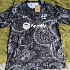 Svart Nike FC Barcelona Dri-FIT tröja XL - Snygg svart FC Barcelona fotbollströja från Nike med Dri-FIT-material. Tröjan har ett unikt paisleymönster i grått och vitt, korta ärmar och rund hals. Officiella klubbmärket och sponsorlogga på bröstet. Perfekt för dig som vill sticka ut på planen eller läktaren.