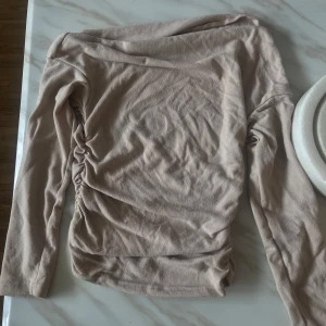 Beige långärmad omlottopp - Trendig beige topp med lång ärm och snygg omlottdetalj på sidan som ger en cool rynkad effekt. Perfekt för dig som gillar stilrena plagg med twist. Mjuk och skön känsla, passar till både jeans och kjol.