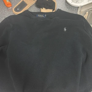 Svart stickad tröja Polo Ralph Lauren - Svart stickad tröja från Polo Ralph Lauren i storlek M. Klassisk design med rund hals och broderad logga i vitt på bröstet. Tröjan har långärmade ärmar och ribbade muddar vid hals, ärmslut och nederkant. Perfekt för dig som gillar stilrena och tidlösa plagg.