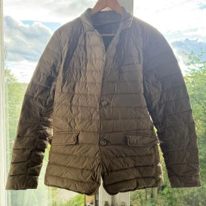 Brun quiltad dunjacka Massimo Dutti - Snygg brun quiltad dunjacka från Massimo Dutti med klassisk krage och två framfickor med lock. Jackan har knappstängning framtill och är tillverkad i dun-material som gör den lätt och varm. Perfekt för hösten. !! Litet brännhål på midjan !!